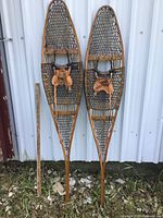 Pair of wooden framed snowshoes with tightly woven webbing and leather bindings, placed outdoors against a metal siding wall with a measuring stick on left for scale.