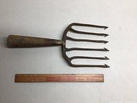 Photo showing one iron farm pitchfork head with five pointed tines lying beside a 12-inch ruler for scale; the pitchfork is rusty and used.