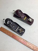 Two Stanley block planes on a white surface with a wooden ruler for scale. One plane is black, the other maroon with visible Stanley label.