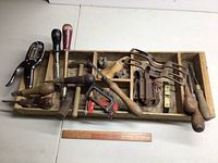 Full wide image of assorted vintage hand tools in a divided wooden box, seen from above, showing metal and wooden tools including hammers, drills, and clamps.