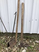 Photo showing both the waffle iron with its square cast iron plates and long wrought handle alongside the wooden handled shipwright's adze standing upright on a gravel surface.