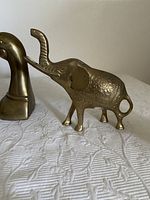 Photo showing brass duck head figurine and brass elephant figurine side by side on white textured cloth.