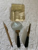 Vintage desk set components laid out on a textured white surface including magnifying glass, letter opener, gold-toned pen, and worn fabric case