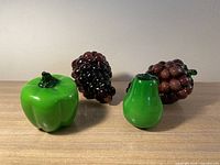 Four glass fruit figures: two purple grape bunches, one green pear, one green bell pepper on wooden surface.