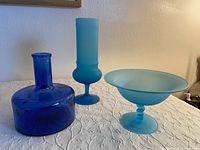 Front view of three blue glass items: cobalt blue bottle, frosted tall glass, and frosted pedestal bowl.