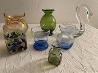 Lot of seven assorted small glass pieces on a white patterned tablecloth including an owl-shaped glass, green vase, swan figurine, and others.