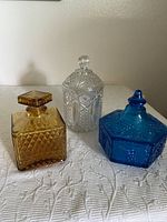 Three glass containers on a white textured cloth: clear cut glass decanter in back, amber square jar at left, blue hexagonal jar at right.