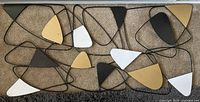 Abstract metal geometric wall hanging showing various triangular shapes in black, white, gold, and bronze connected with black metal lines placed on a carpeted floor.