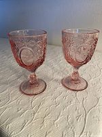 Two pink glass goblets with embossed floral and oval medallion designs on a textured surface, placed on a white patterned cloth.