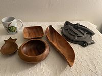 Four Monkey Pod wooden dishes (small round bowl, leaf-shaped dish, square shallow dish, pineapple-shaped plate) and one cast iron tortilla press placed on beige textured fabric.