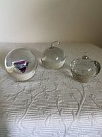 Three glass paperweights on white textured surface. Round with purple swirl, clear apple shape, and clear bubble apple shape.