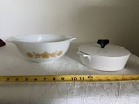 Photo of the two dishes side by side on a white textured surface with a tape measure showing about 12 inches width, showing the Pyrex dish with gold pattern and the white Corning Ware server with black knob lid.