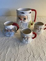 Front view of Santa face pitcher and three Santa mugs on a white textured cloth surface, showing detailed facial features and red handles
