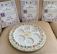 Photo showing 3 Valley Beth Shalom Haggadah booklets standing behind a large white Passover Seder plate with seven sections and colorful floral border with gold trim.
