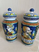 Two ceramic apothecary jars with original lids, shown side by side on a surface. They feature hand-painted figure portraits, bright colors, and decorative bands.