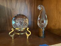 Front view of the clear glass crystal ball paperweight with internal bubbles, resting on a polished brass stand, placed beside the clear flame glass sculpture with a copper base, displayed on a wooden shelf.