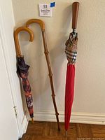 Three items displayed upright against a wall: the Burberry umbrella, floral umbrella, and wooden cane.
