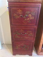 Front view of Chinese style filing cabinet showing three red drawers with scenic mountain landscape artwork in gold and green with metal handles.