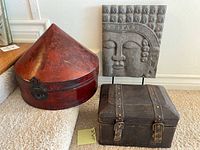 Three items shown: red lacquer hat box, brown leather snap buckle box, and grey resin Buddha sculpture on wood stand