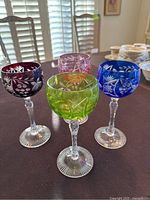 Set of four colored crystal wine glasses in purple, pink, green, and blue with detailed cut patterns on the bowls and clear decorative stems, photographed on table.