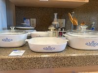 Group photo of three white Corning Ware casserole dishes with blue cornflower pattern and glass lids on two dishes, set on kitchen counter.