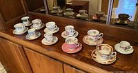 11 different fine bone china teacup and saucer sets displayed in two rows on a wooden surface.