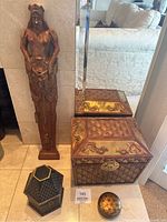 Wide view of tall carved female statue standing next to two larger decorative boxes and smaller hexagonal trinket box with an additional small decorative round dish in foreground, set on tiled floor in front of mirror and sofa.