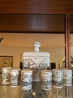 Vintage German porcelain decanter and six small cups displayed on a shelf with detailed landscape artwork