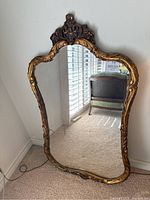 Full view of the large gold gilt mirror leaning against a wall, showing the ornate carved top and gold finish.