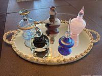 Wide view of four decorative glass perfume bottles on ornate oval mirror base showing various bottle shapes, colors, and stopper designs.