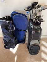 Three bags side by side: black/blue stand bag, blue travel bag, grey bag loaded with clubs.