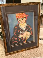 Framed reproduction print of woman in red madras headdress seated in colorful patterned clothing