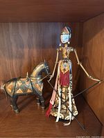 Full view of the Indonesian puppet alongside the horse figurine in a wooden cabinet shelf showing the traditional costume and detailing on both items.