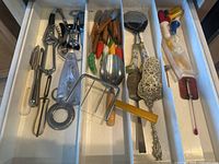 Drawer containing assorted kitchen utensils: ceramic handled pizza slicer, wooden cheese cutters, bakelite cheese slicer, corn cob holders, potato peelers, cake servers, metal kitchen tools