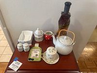 Photo shows Belleek Clover collection including teapot with bamboo handle, hexagonal sugar pot with lid, red polka dot creamer, salt and pepper shakers, olive oil dish, and white dishes with green floral clover pattern on wooden table.