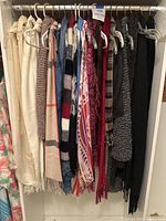 Full view of 12 assorted scarves hanging on a rod showing different colors, materials, and patterns including silks, cotton knits, woven and hand-knit fabric types.