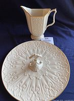 Lenox vintage pitcher with floral relief and gold trim positioned behind Lenox Chateau serving tray with carved pattern and central handle, both white ceramic with gold accents.