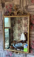 Full view of the rectangular mirror mounted on floral wallpaper, showing the intricate gold ornate frame with filigree decorations on top and bottom.