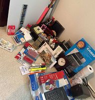 Full view of the lot showing Boston electric pencil sharpener, boxed Casio printing calculator, Canon printing calculator, writing utensils, receipt paper, photo paper, tape dispenser, playing cards, and other items.