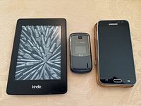 Front view of Kindle e-reader, Verizon LG flip phone, and Samsung Galaxy J3v smartphone lying flat on a surface.