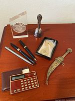 Silver bell, acrylic peace medallion, decorative brass cutting utensil inscribed 'Istanbul', three pens, vintage calculator with leather case displayed on wood table.