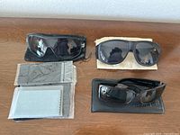 Three pairs of black frame sunglasses on a wooden surface with respective cases and lens cleaning cloths arranged nearby.
