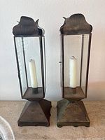 Front view of two tall brass lantern candle holders with white candles inside, showing aged brass and glass panes.