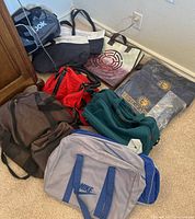 Overview of assortment of totes and duffel bags on carpeted floor near wooden furniture and wall outlet