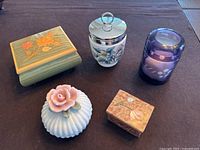 Five trinket boxes: green wood box with painted flowers, ceramic jar with floral painting and silver rim, purple glass decorated jar, white ceramic jar with pink rose on lid, small rectangular wooden box with bird design.