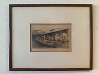 Photo of the framed etching hanging on a white wall showing the entire piece with brown wooden frame and cream mat.