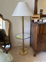 Full view of vintage brass floor lamp with round glass table and white lampshade, placed next to wooden chest and a mirror.