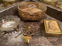 Glass-lidded jewelry/trinket box with gold metal intricate filigree base, brass cherub towel holder, and gold with green trinket box on countertop.