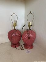 Pair of vintage rose blush ceramic shell-shaped table lamps with brass fittings and no lampshades, placed side-by-side on a flat surface.