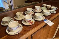 Photo showing 8 matching teacup and saucer sets with floral patterns and various colors arranged on a wooden cabinet.